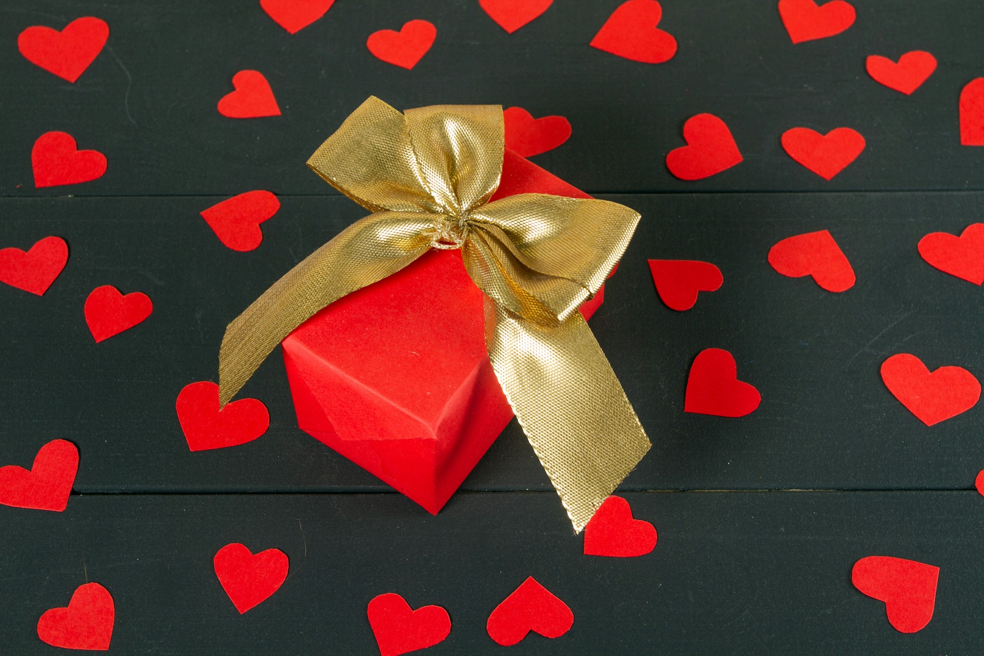 picture of red gift box with gold bow on top surrounded by red paper hearts on black paper