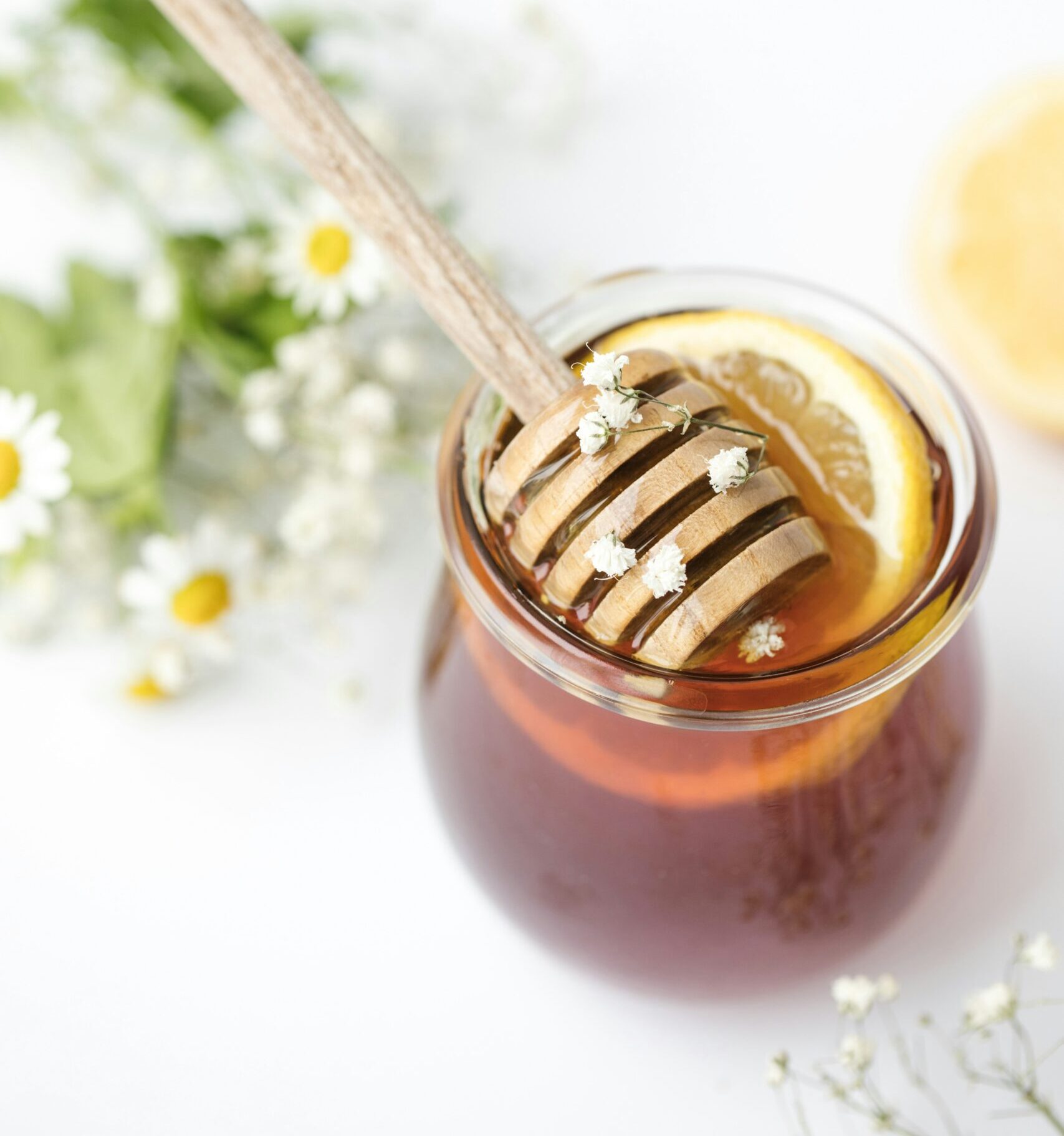 Photo of honey pot with stirrer and lemon slice in it. Out of focus, in the background are small white flowers with yellow centers and some baby's breath flowers.