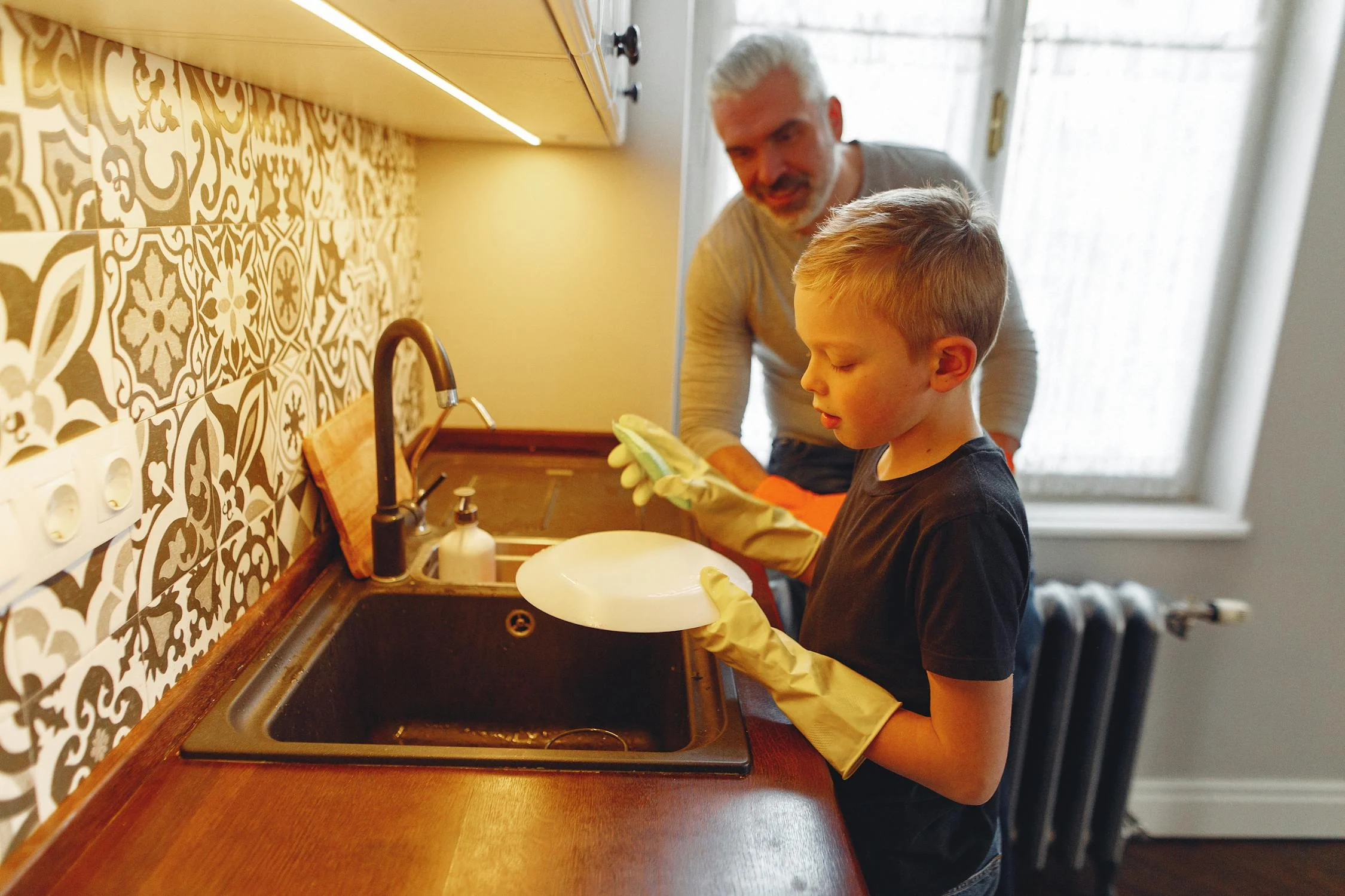 Blond haired boy washes a white dish as father watches and smiles