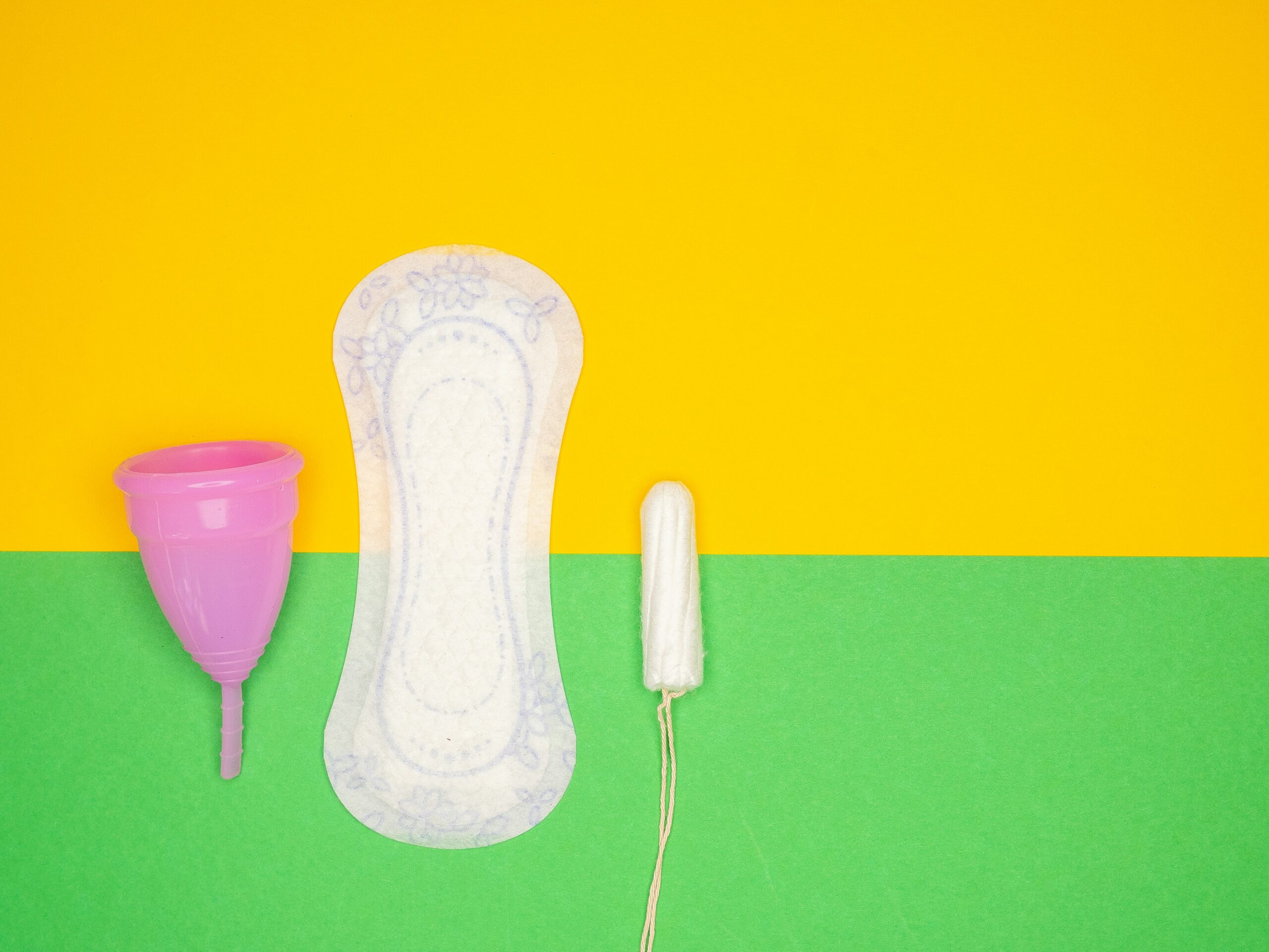 Photo of a half yellow, half green background with a menstrual cup, a pad and a tampon placed on the lower left-hand side