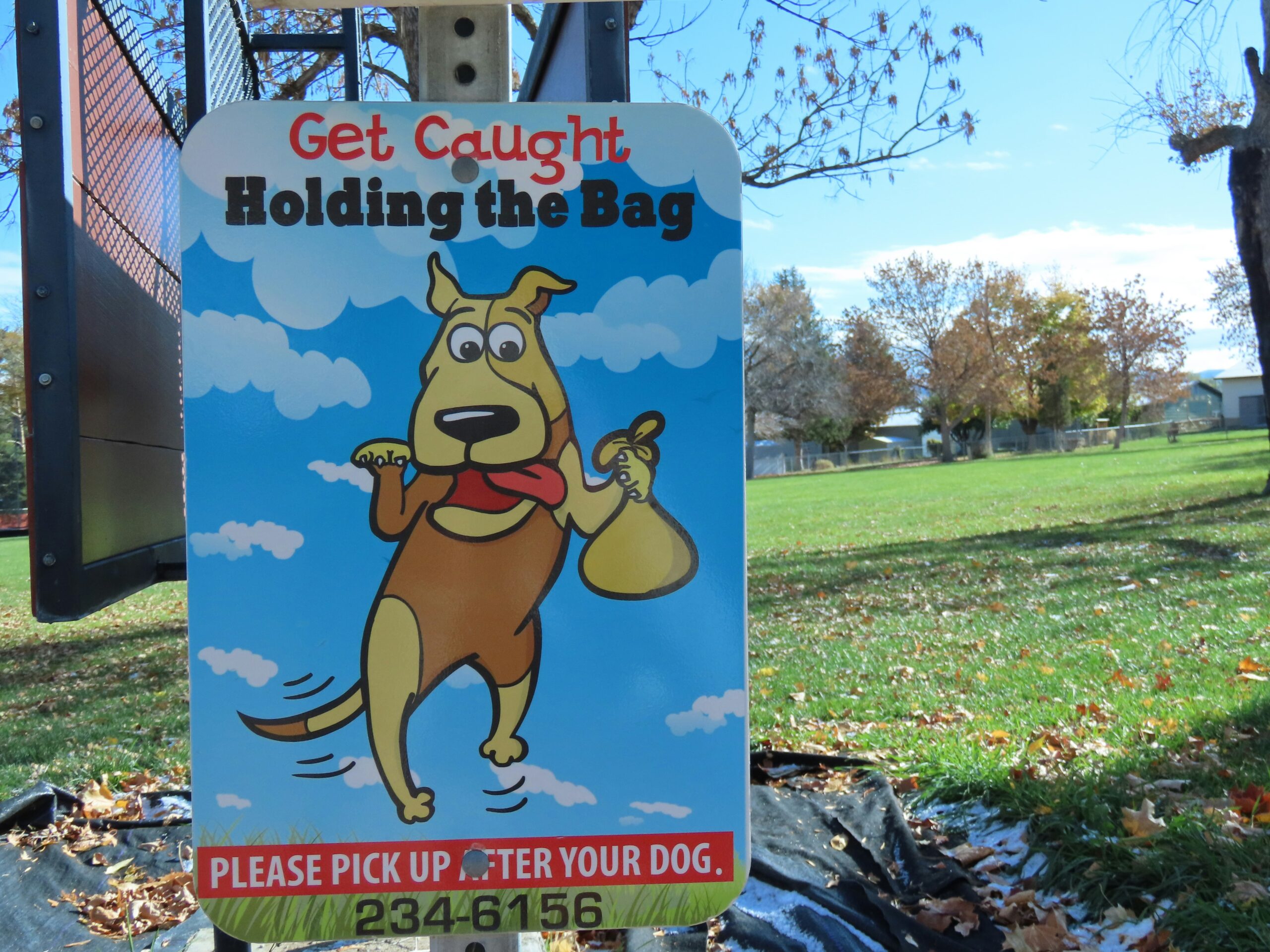 photo of a sign. on the sign is a dog holding a poop bag. the sign asks dog owners to clean up after their pets.