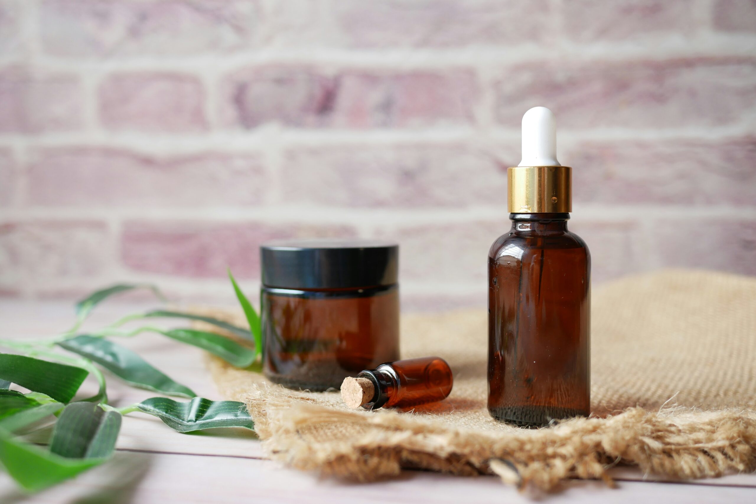 Photo of three brown glass vials on top of a light brown fabric with green leaves scattered around. The background is a brick wall.