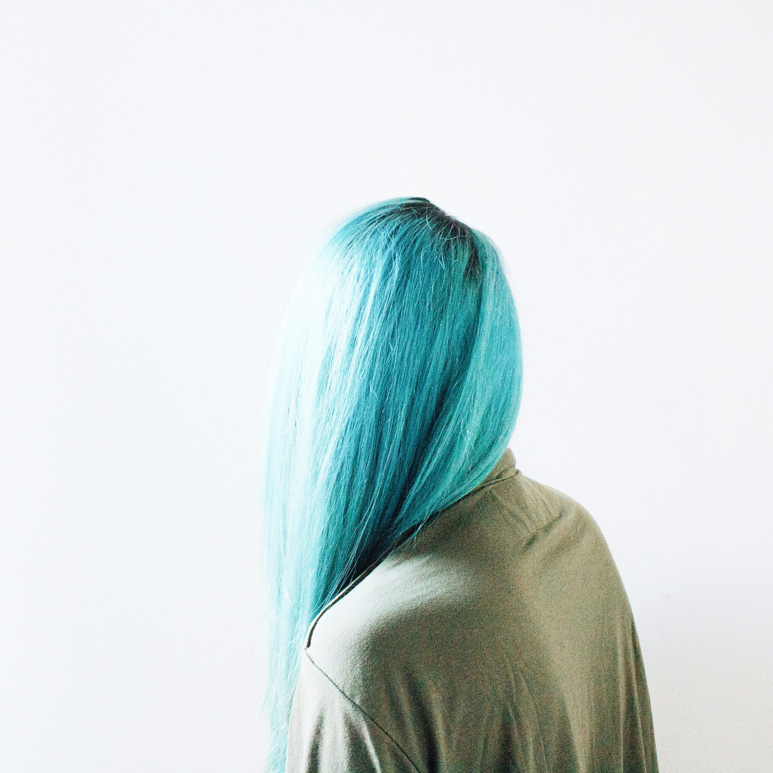 Photo of person with long, blue hair facing away from the camera and wearing an olive green shirt. White background.