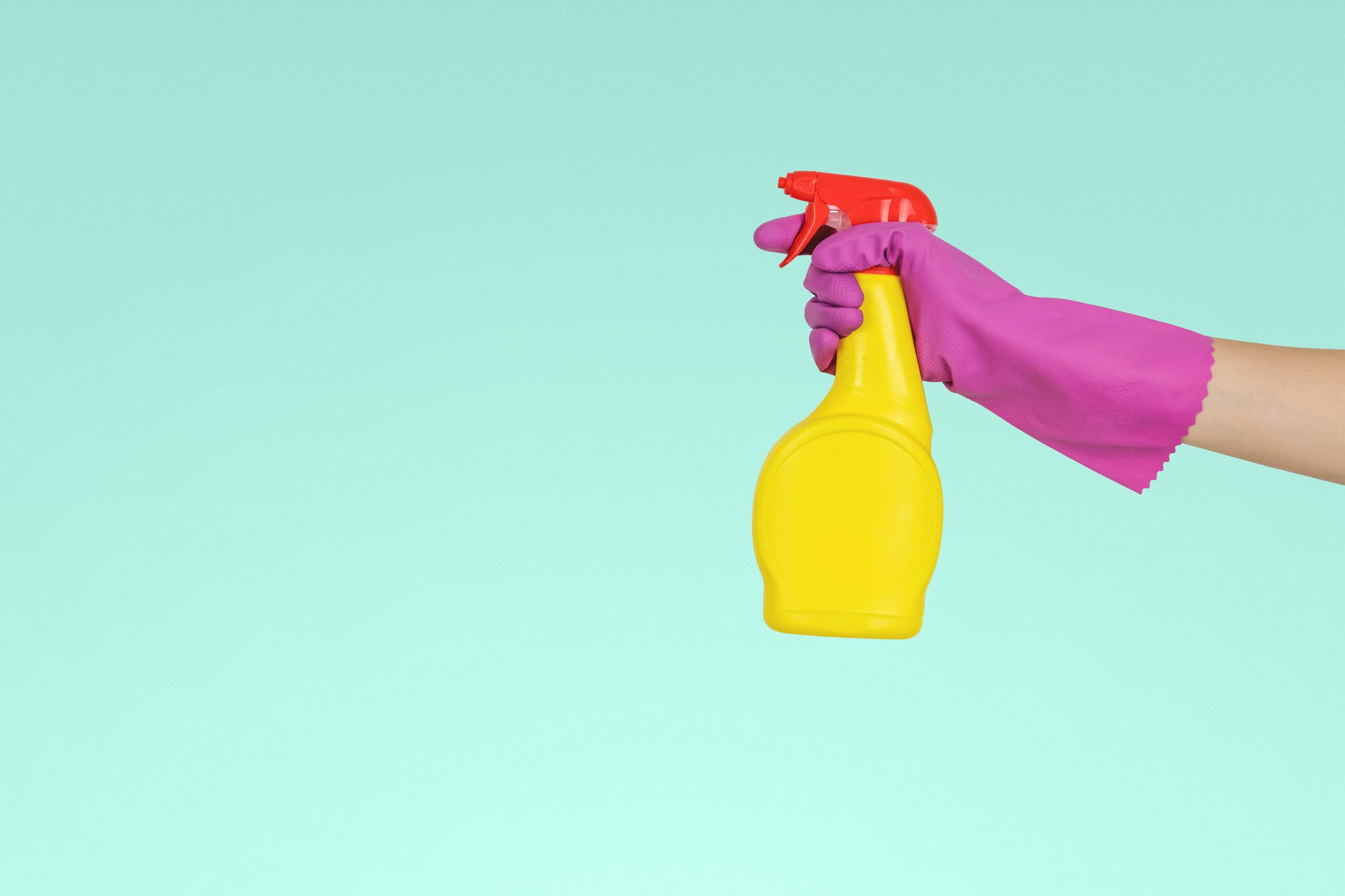 Arm covered in a pink dish washing glove, holding a yellow spray bottle in front of a light blue background