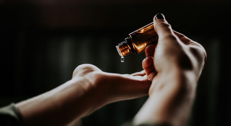 photo of two hands, one holding a small glass amber dropper and dropping essential oil onto the palm of the other hand. black background