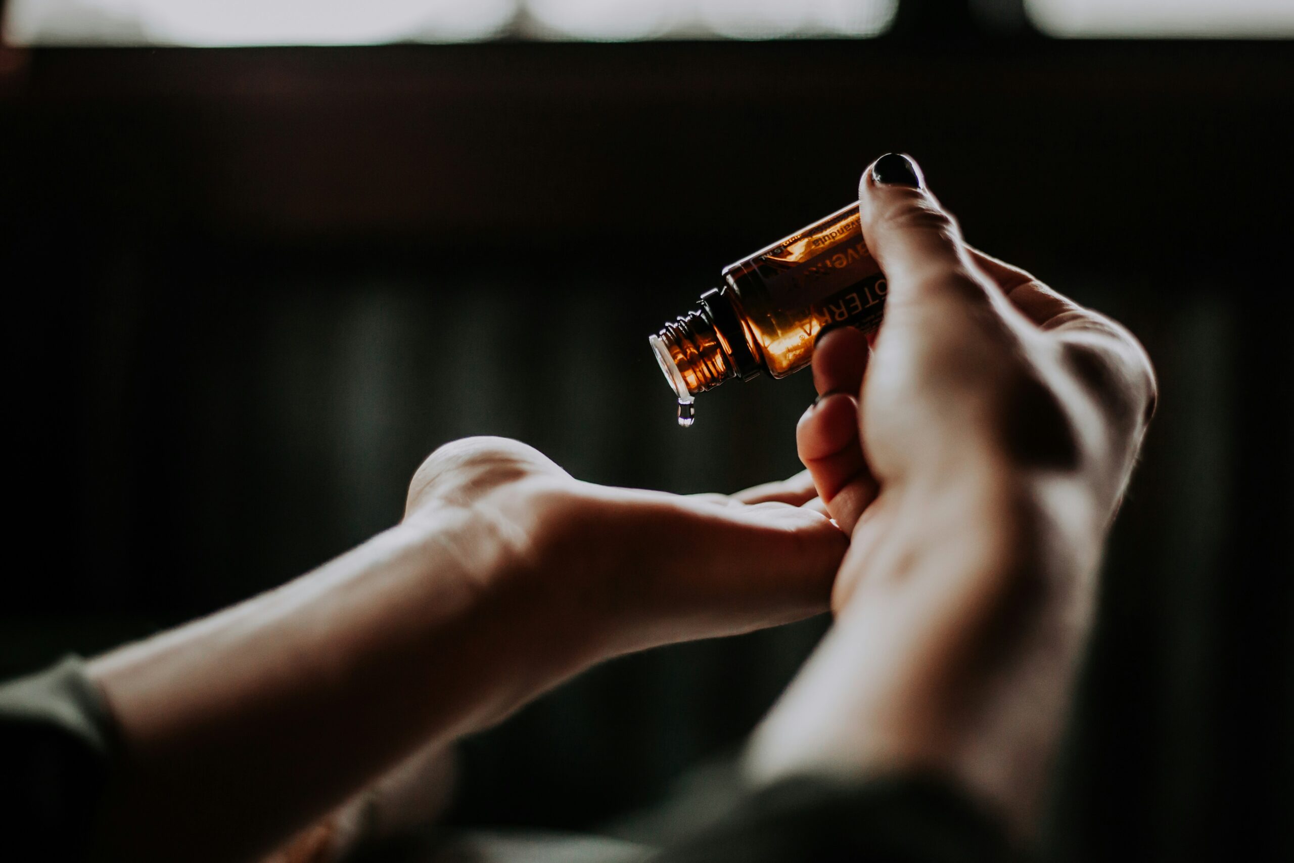 photo of two hands, one holding a small glass amber dropper and dropping essential oil onto the palm of the other hand. black background