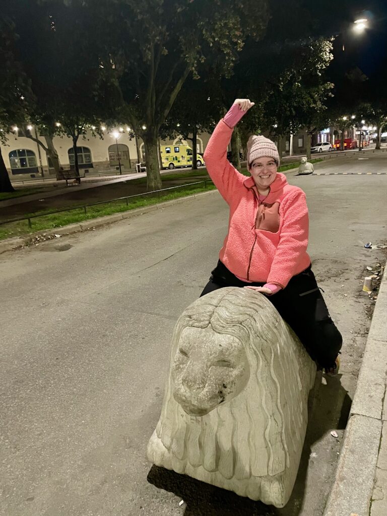 Woman wearing a pink sweater and black pants while pretending to ride a stone lion like a mechanical bull.