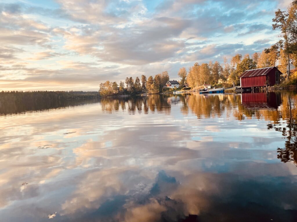 Photo of landscape. Lake. Reflected in the water is the clouds, a red house and tall fall-colored trees.