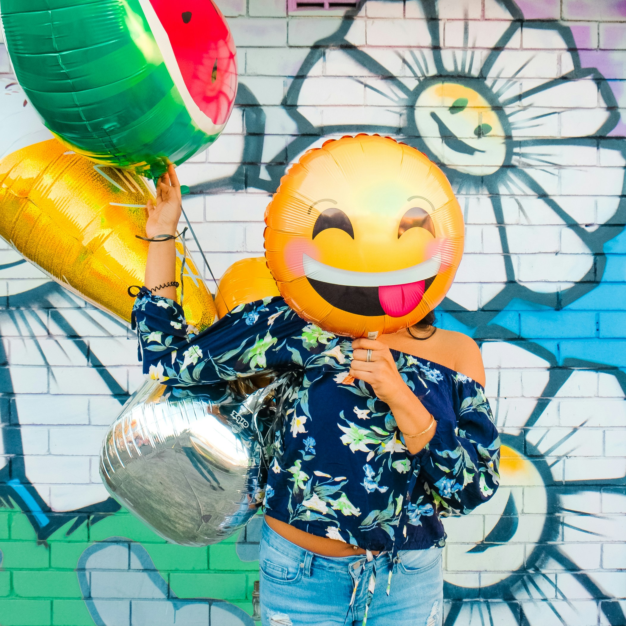 Woman in a floral blue blouse holding a happy face balloon over her face in front of a colorful brick wall with gratified daisies.