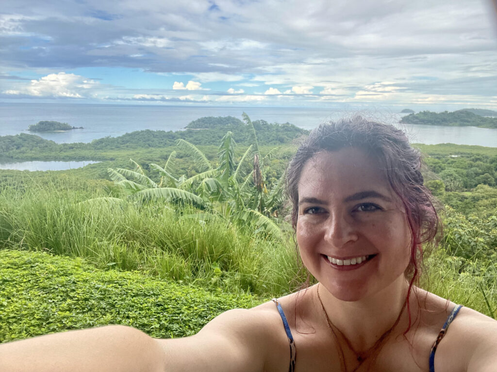 Selfie of woman with wavy, red hair. In the near background is green jungle. In the distant background is the ocean. Islands are visible. The sky is blue and full of white clouds.