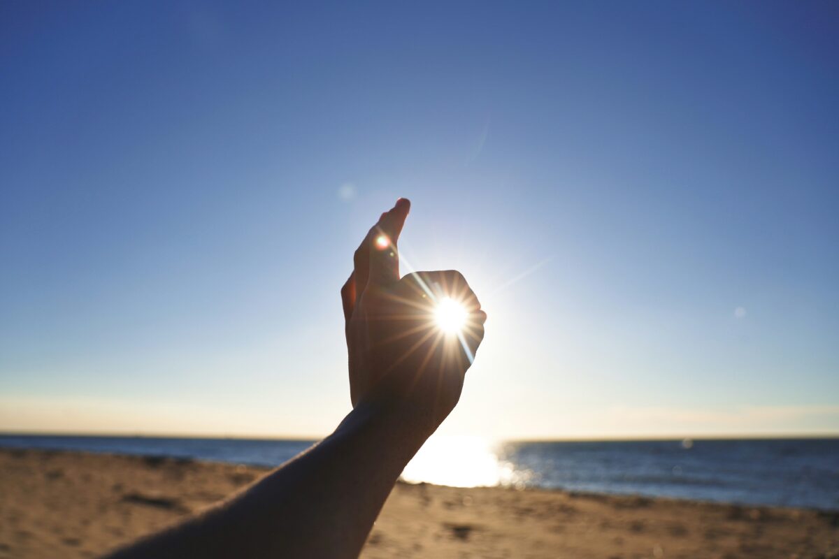 Person raising left hand in an OK symbol over the sun near the sea.